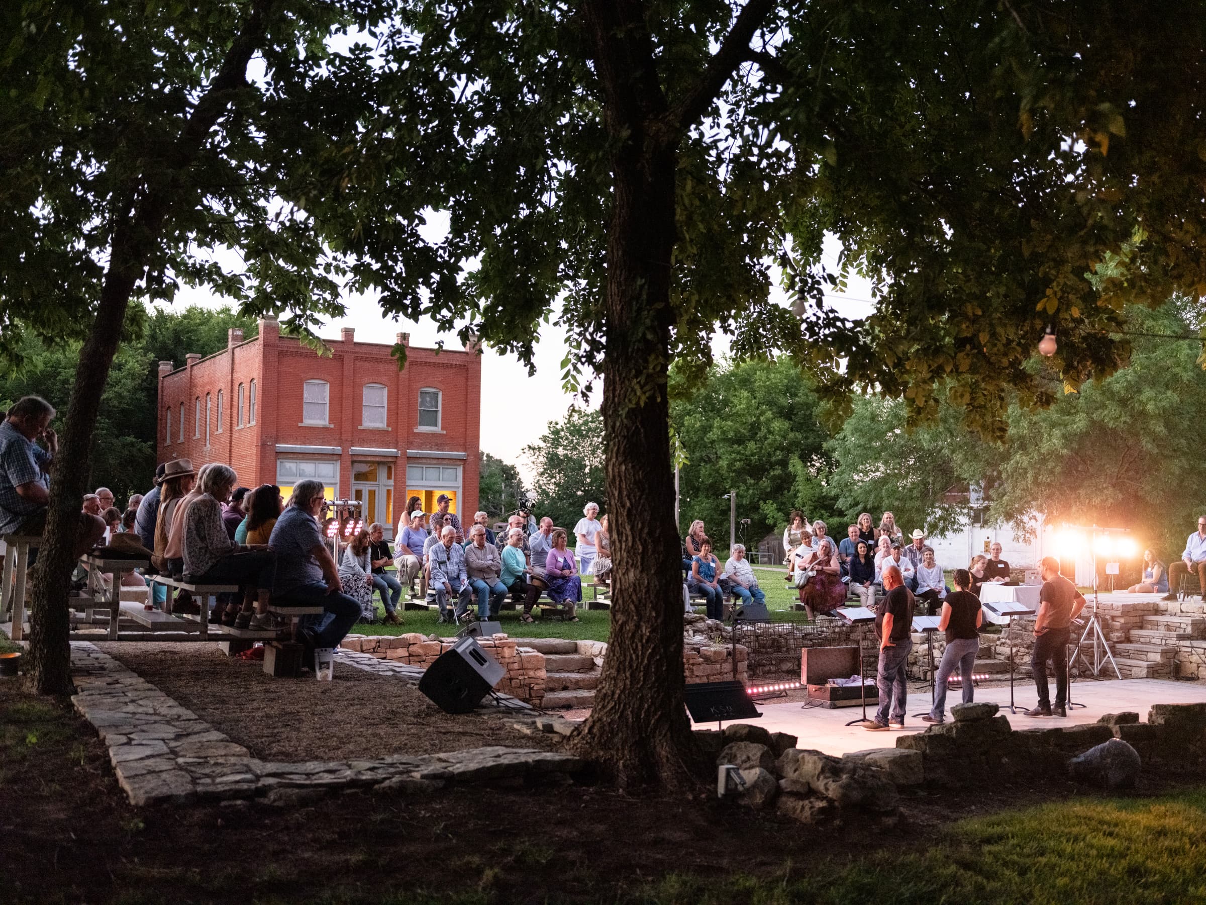 A crowd gathers in a park for an outdoor performance, illuminated by stage lights, with a historic building in the background.