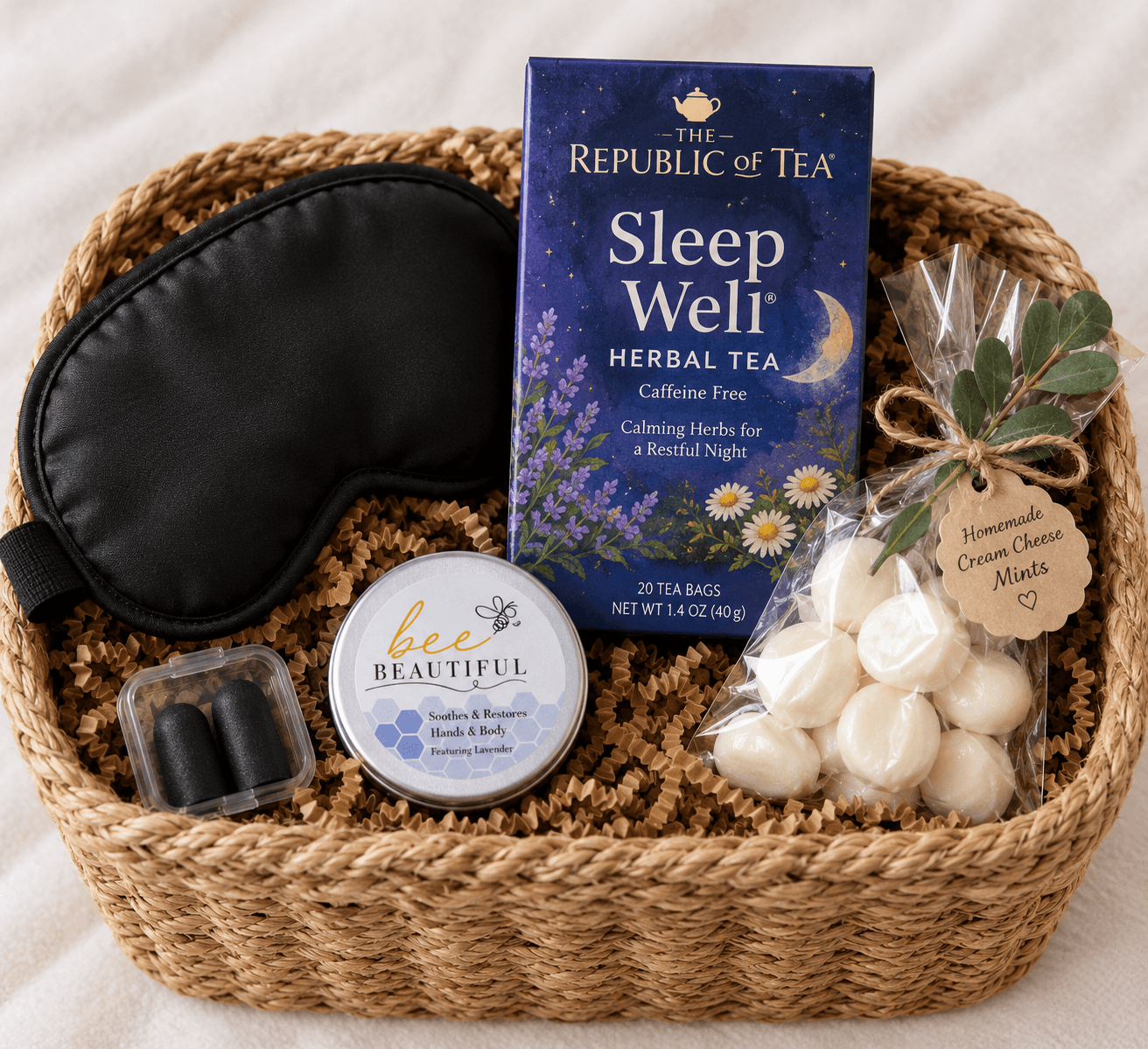A woven basket containing a sleep mask, herbal tea, skincare balm, and homemade cream cheese mints.