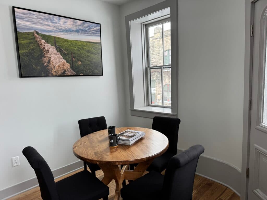A round wooden table with four black chairs, books, and a coffee cup, beside a window and a wall featuring a landscape photo. A round wooden table with four black chairs, books, and a coffee cup, beside a window and a wall featuring a landscape photo.