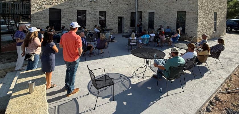 A group of people gathers in an outdoor seating area near a stone building.