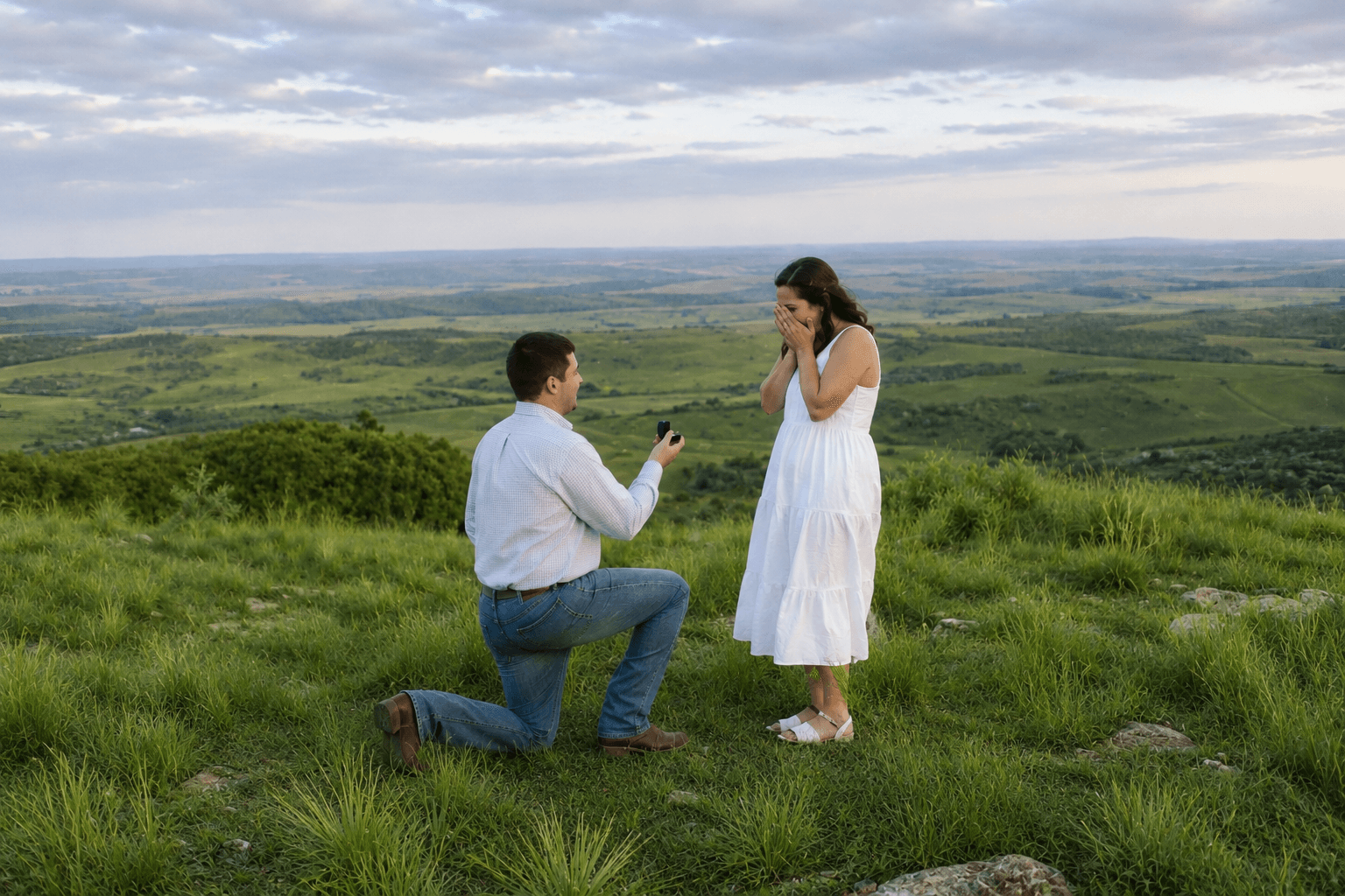A man kneels with an engagement ring while a woman in a white dress reacts with joy on a scenic hillside.