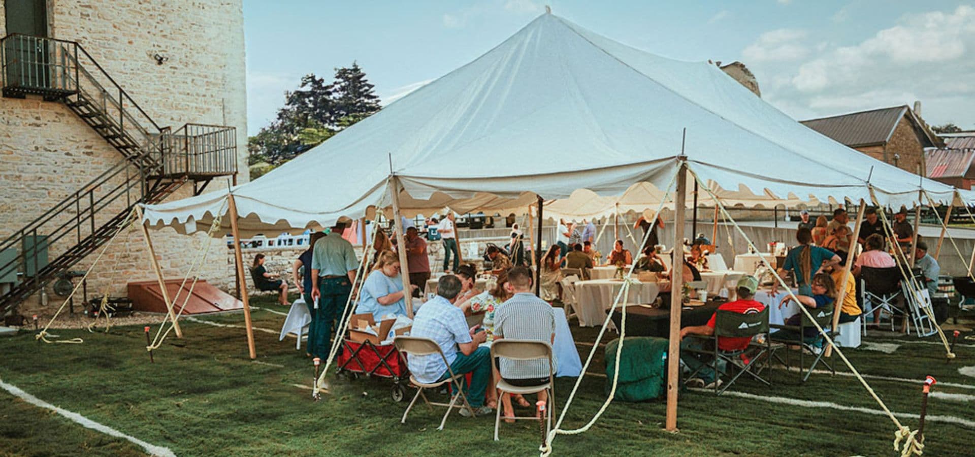 A large gathering under a tent with people sitting around tables enjoying a meal.