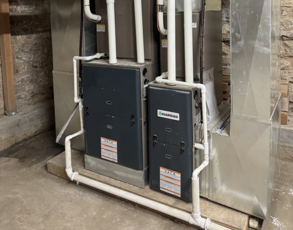 Two black heating units with white pipes, installed in a basement space.