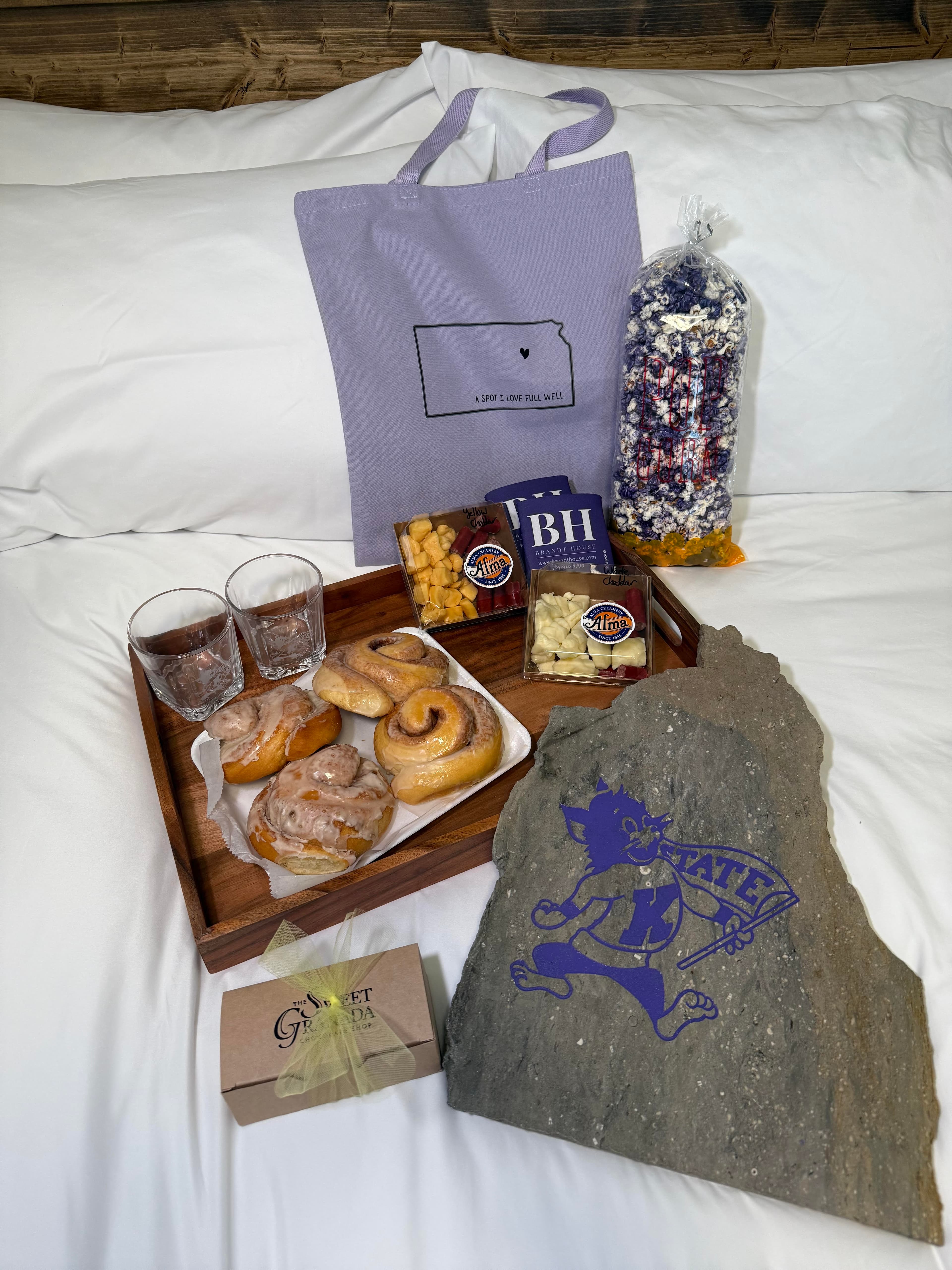A wooden tray with assorted pastries, snacks, and drinks, accompanied by a tote bag and a decorative stone, rests on a bed with white linens.