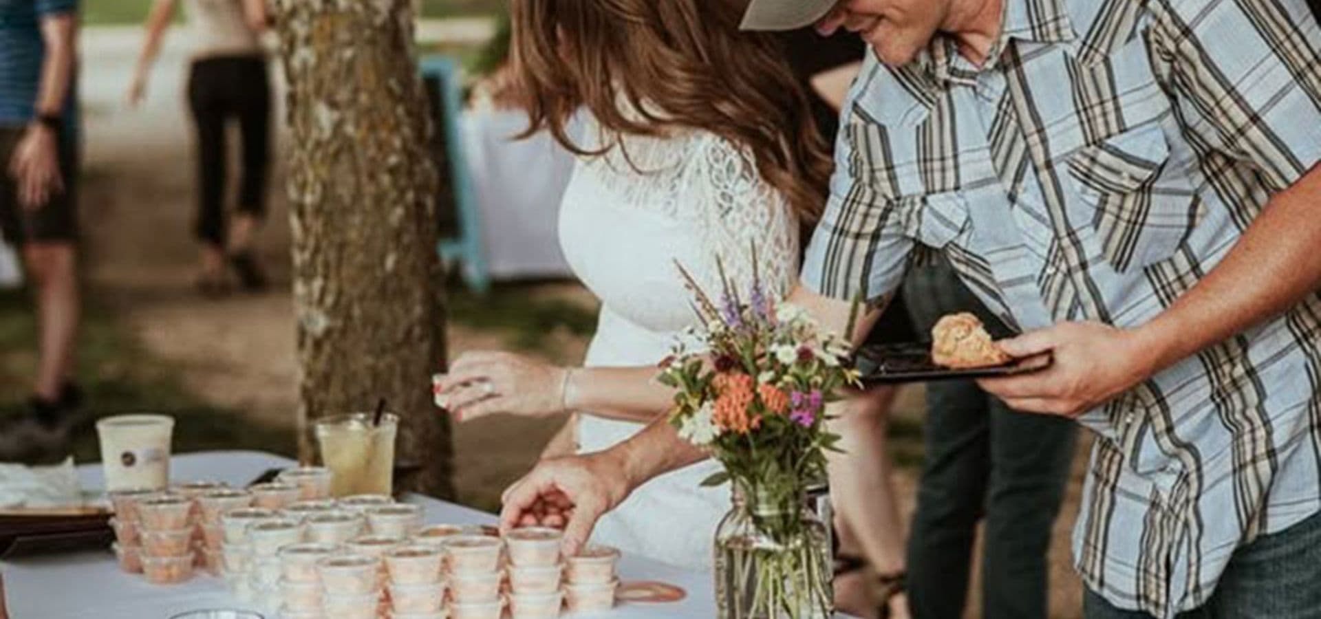 A woman in a white dress and a man in a plaid shirt share a buffet table filled with desserts and drinks, surrounded by greenery.