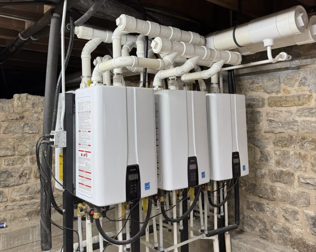 A row of three white tankless water heaters connected with piping in a basement.