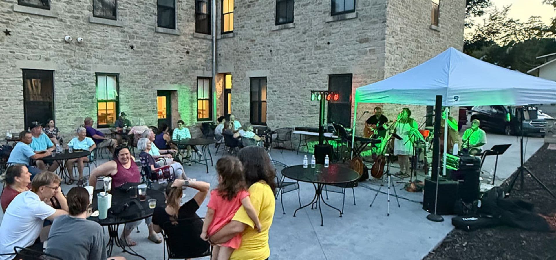 A lively outdoor gathering features a band performing under a tent while people enjoy drinks and conversation at tables.