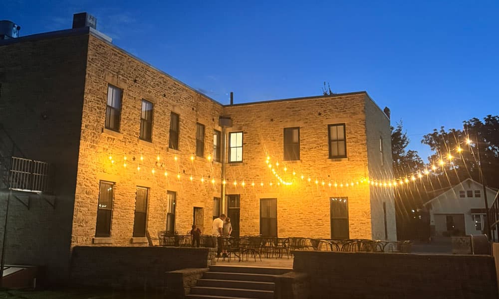 A stone building decorated with warm string lights at dusk, with people gathered outside.
