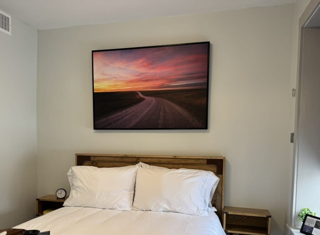 A cozy bedroom featuring a wooden bed, white bedding, and a landscape painting of a winding road at sunset on the wall.