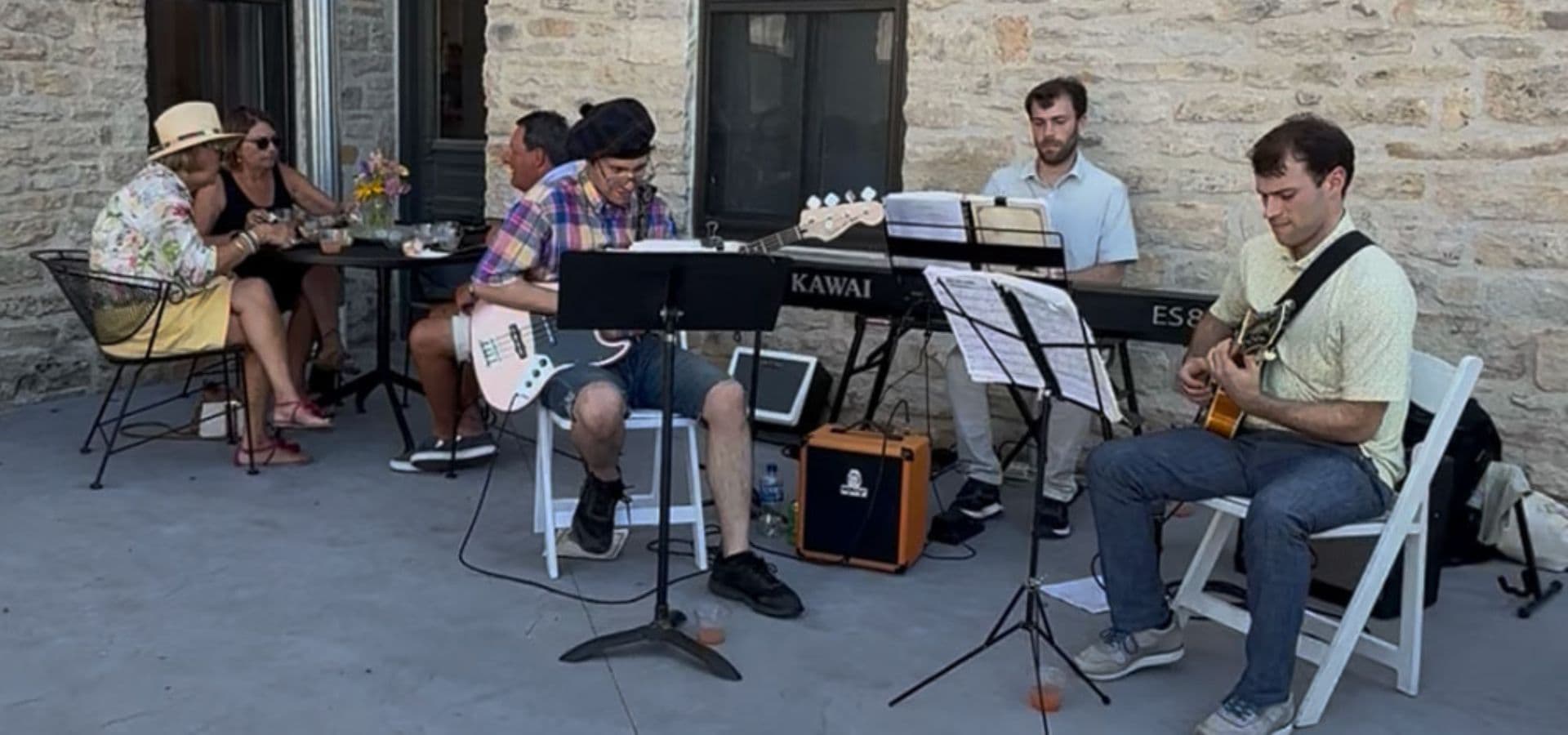 Musicians perform with guitars and a keyboard while people enjoy drinks at an outdoor café.