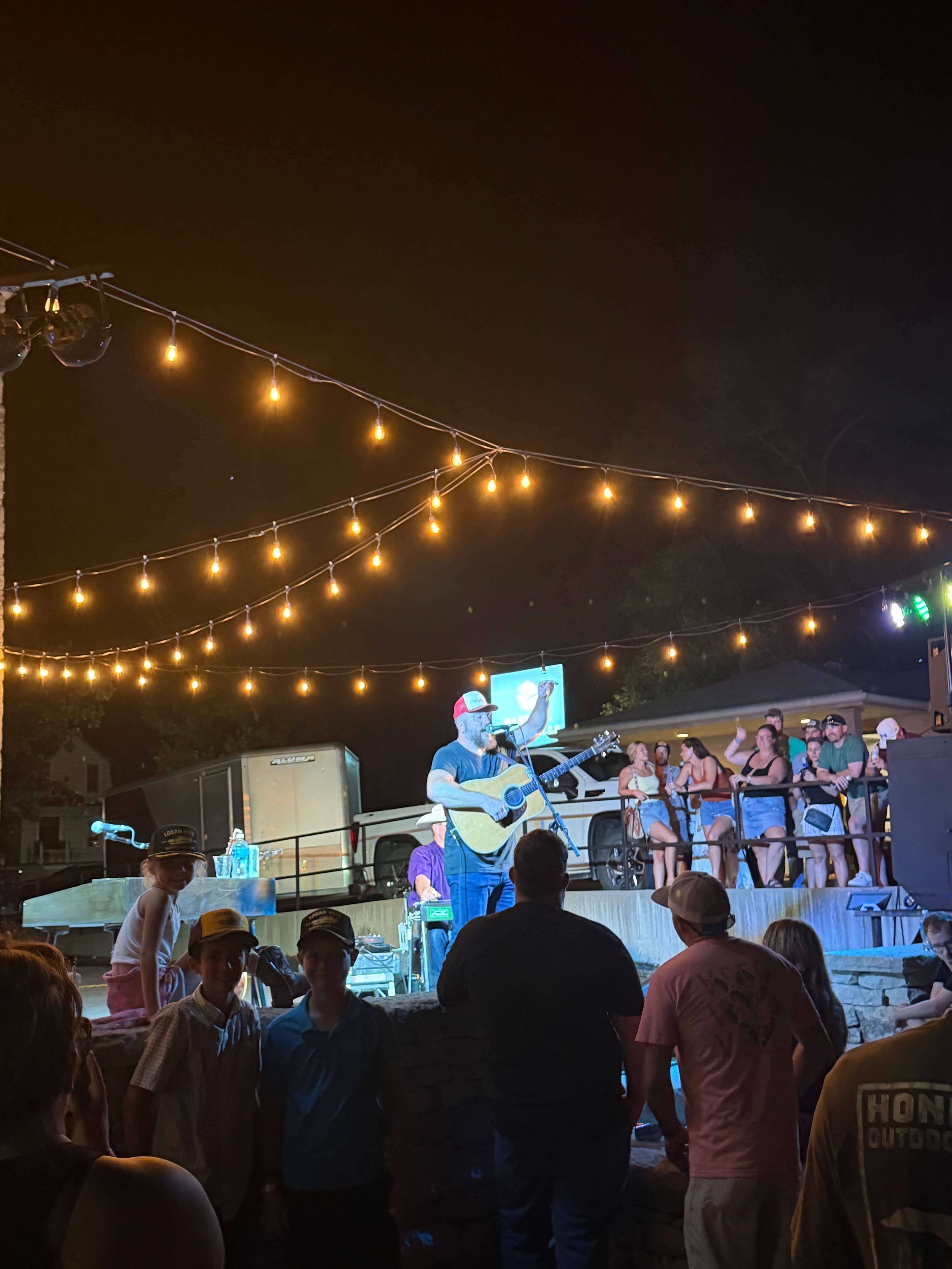 A singer performs with a guitar on stage while an audience watches under string lights at night.