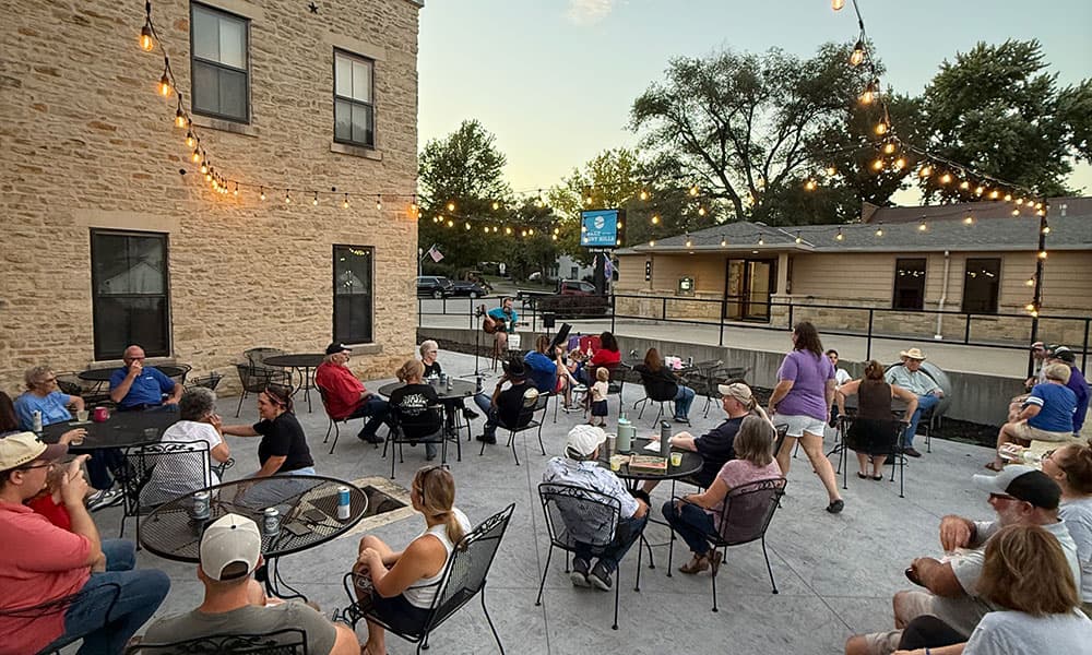 A lively outdoor gathering at dusk, with people seated at tables near a stone building, illuminated by string lights.