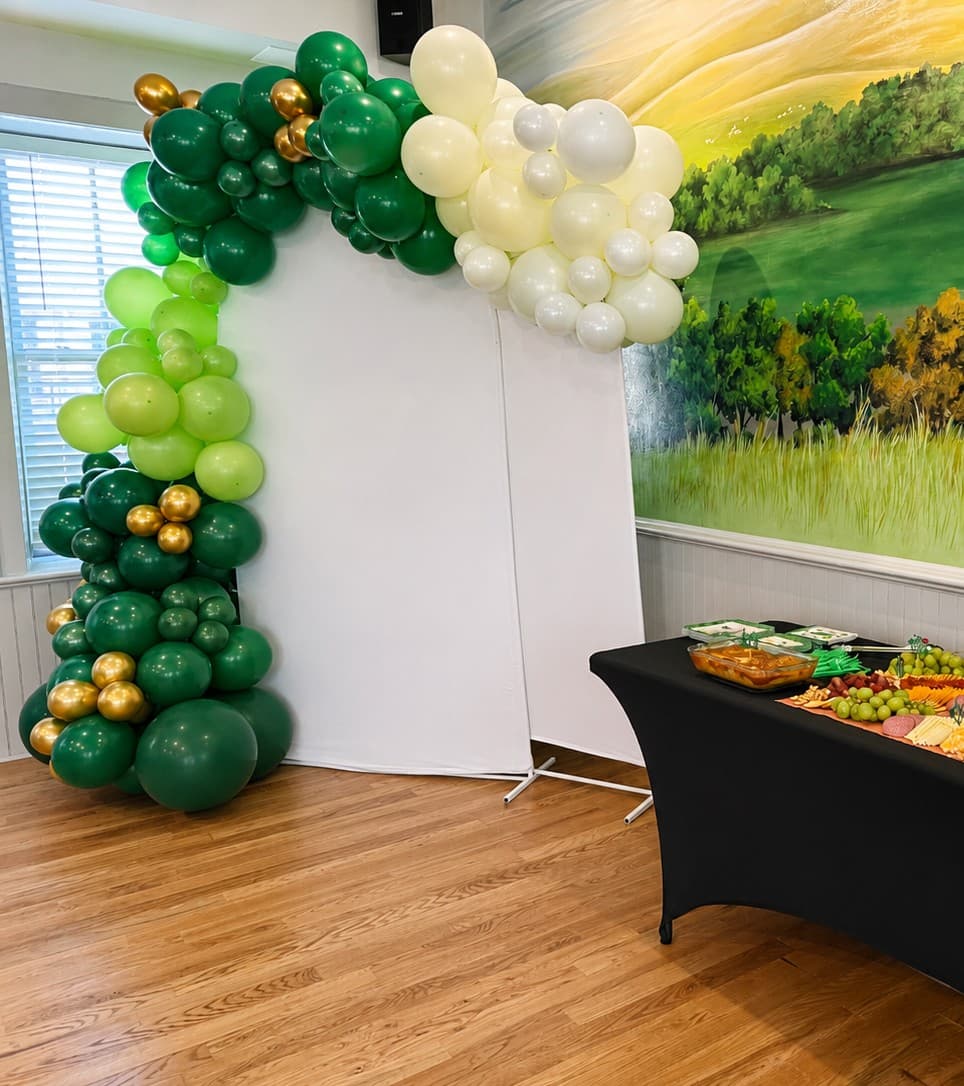 A colorful balloon arch in green, gold, and white frames a plain backdrop next to a table with food and snacks.