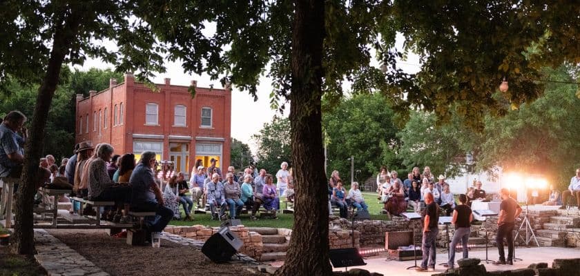 A musical performance takes place in a park with an audience gathered around as night falls.