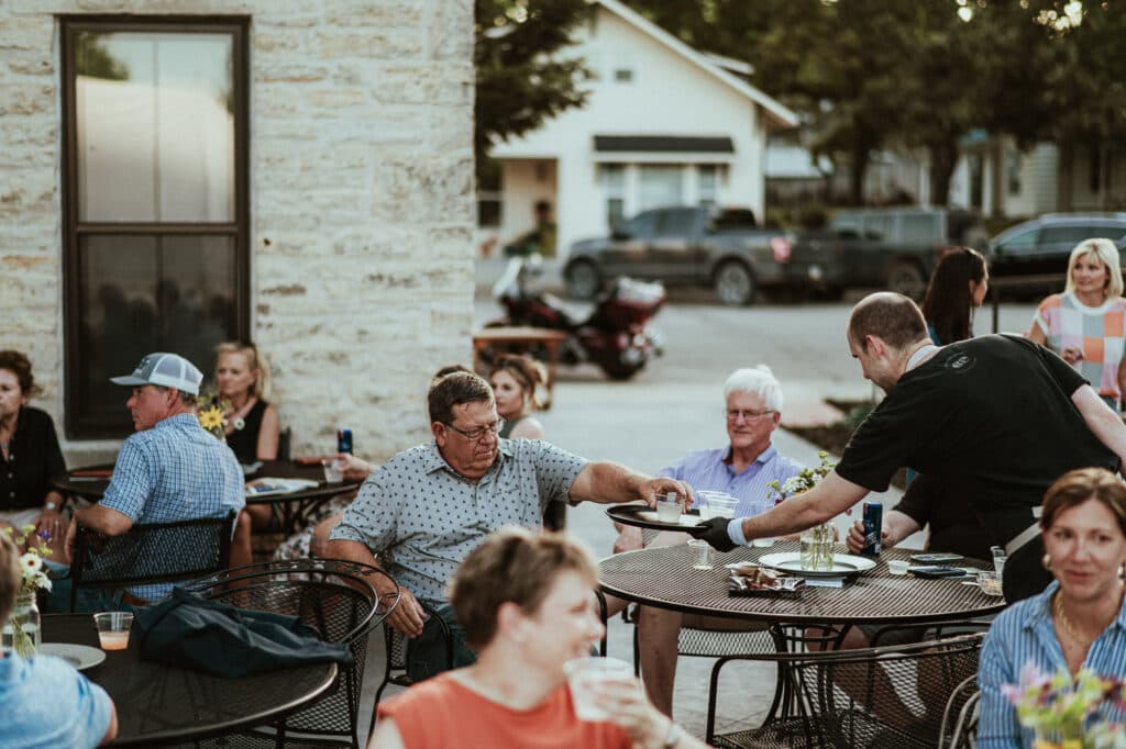 A waiter serves food to a seated guest at an outdoor gathering with people chatting at tables.