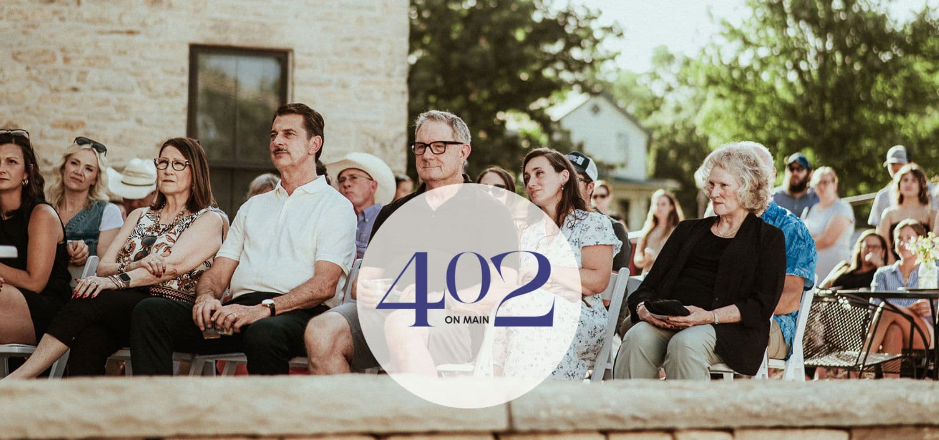 A diverse audience seated outdoors, attentively watching an event, with a prominent logo overlay reading "402 ON MAIN."