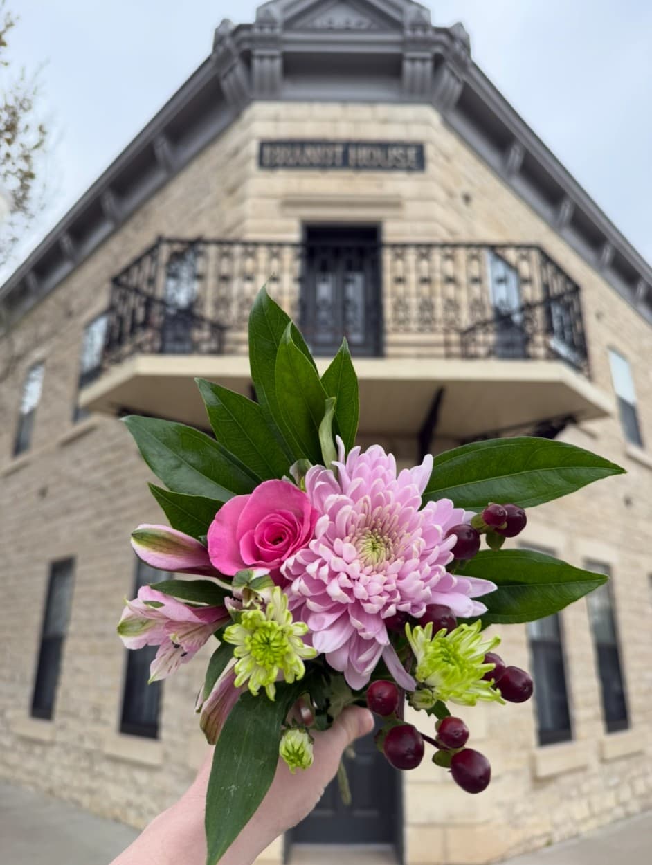A hand holds a vibrant bouquet of flowers against the backdrop of the historic Braundt House.
