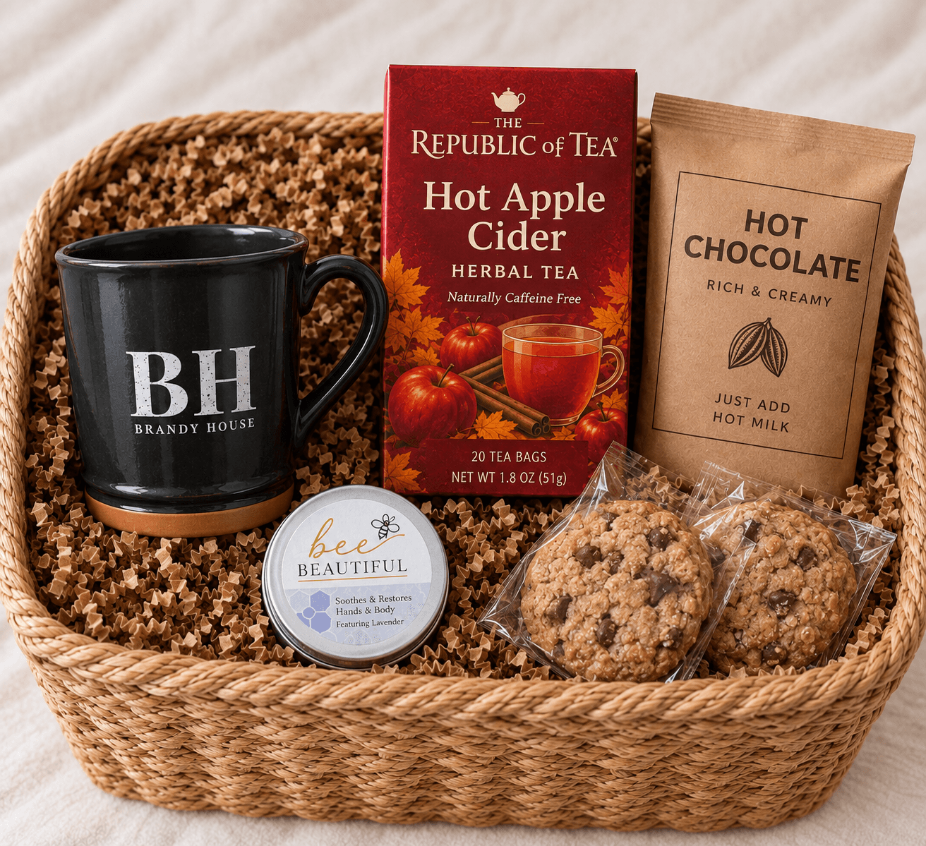 A woven basket contains a black mug, boxes of hot apple cider and hot chocolate, a tin of lotion, and chocolate chip cookies.