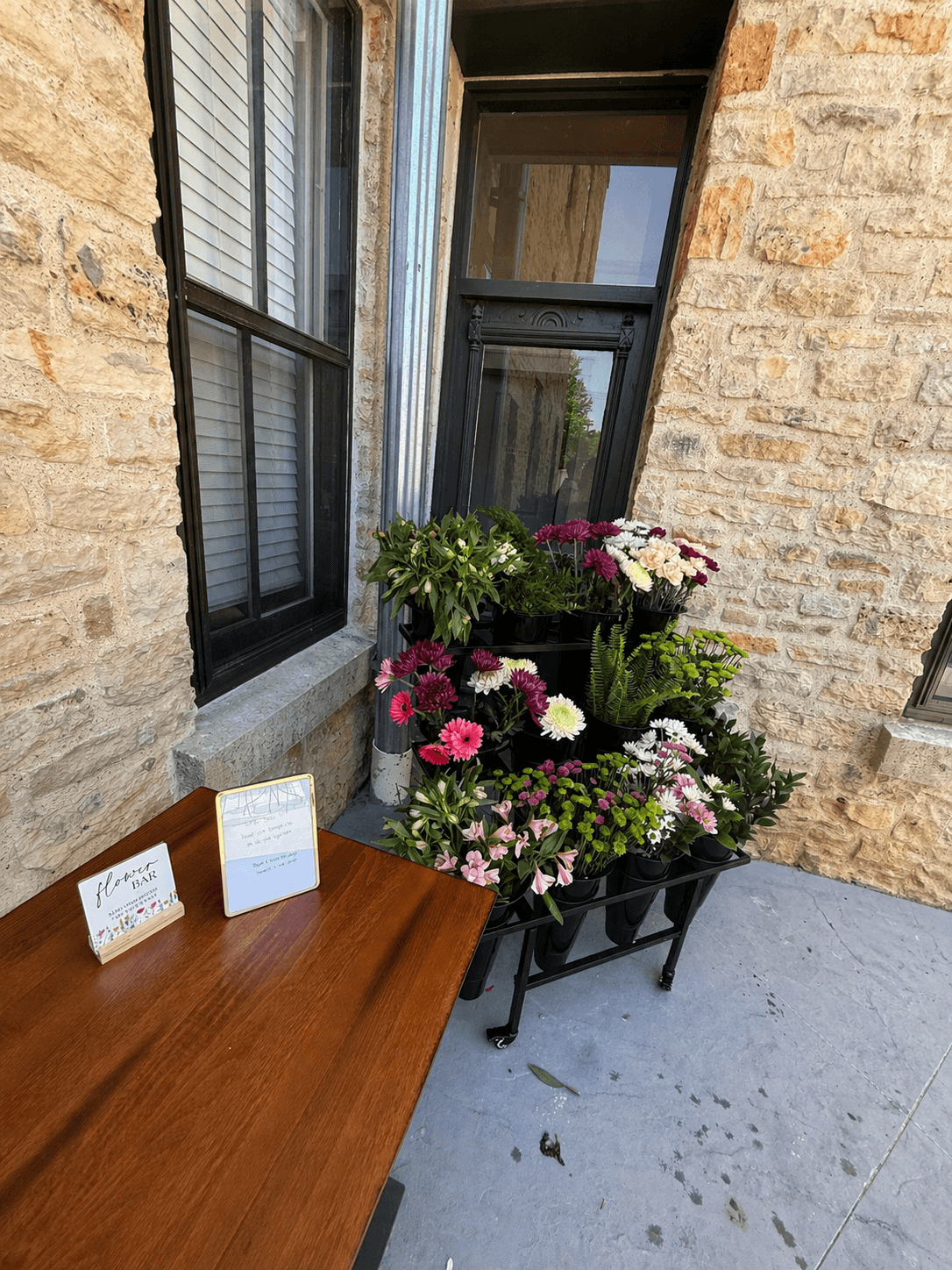 A wooden table with two small signs beside a colorful display of potted flowers on a porch next to a stone wall.