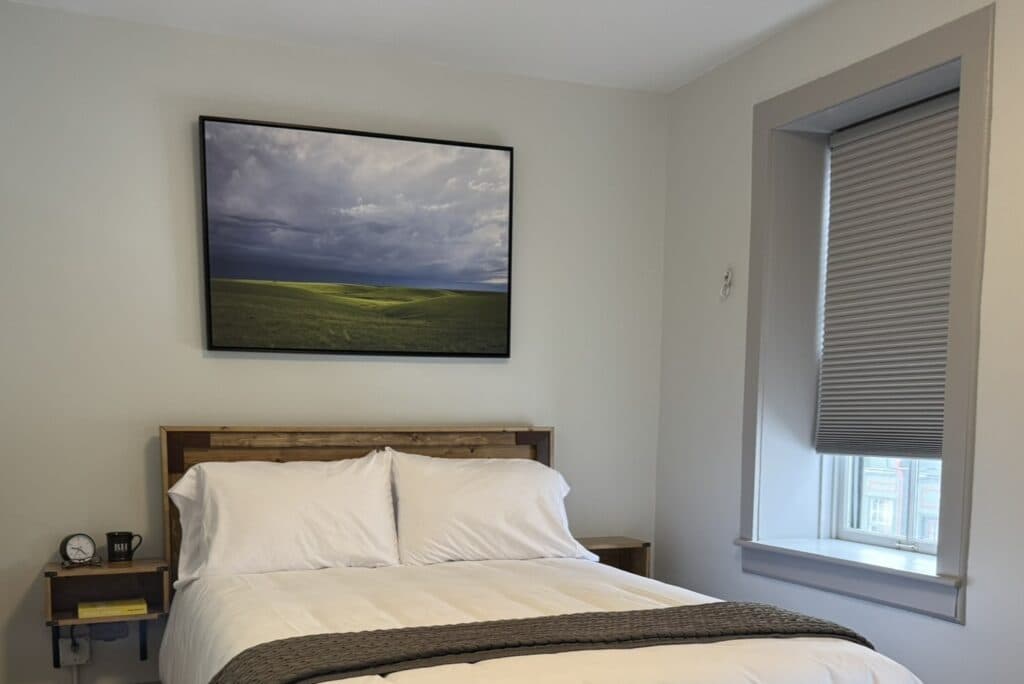 A cozy bedroom featuring a wooden bed, white bedding, and a landscape painting above, with a window and bedside table.