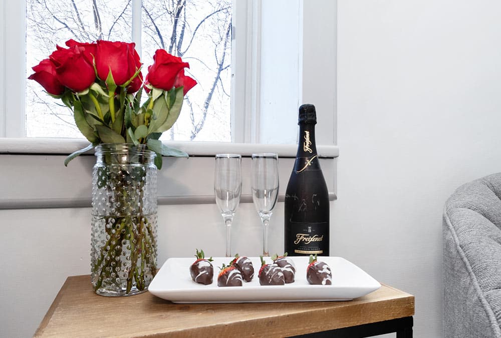 A vase of red roses, two champagne flutes, a bottle of sparkling wine, and chocolate-covered strawberries on a wooden table.