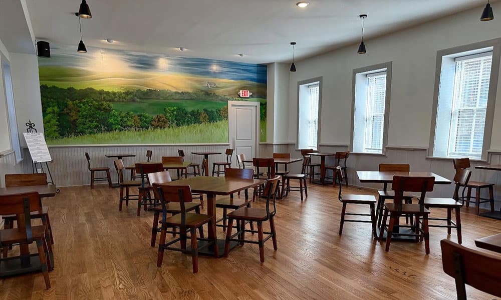 A bright, empty dining room with wooden tables and chairs, featuring a large landscape mural on one wall.