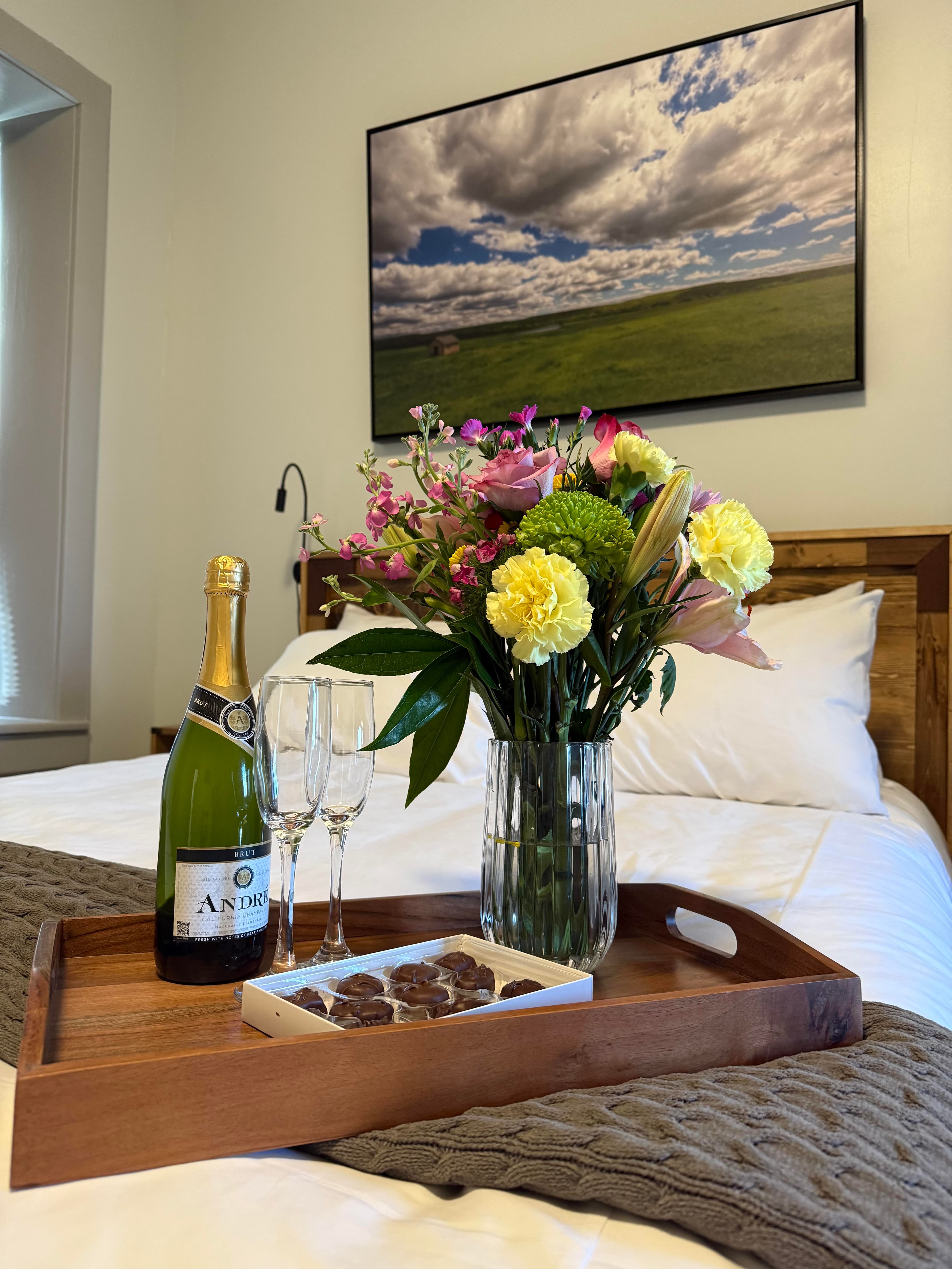 A cozy bedroom setup featuring a tray with champagne, two glasses, a vase of colorful flowers, and a box of chocolates.