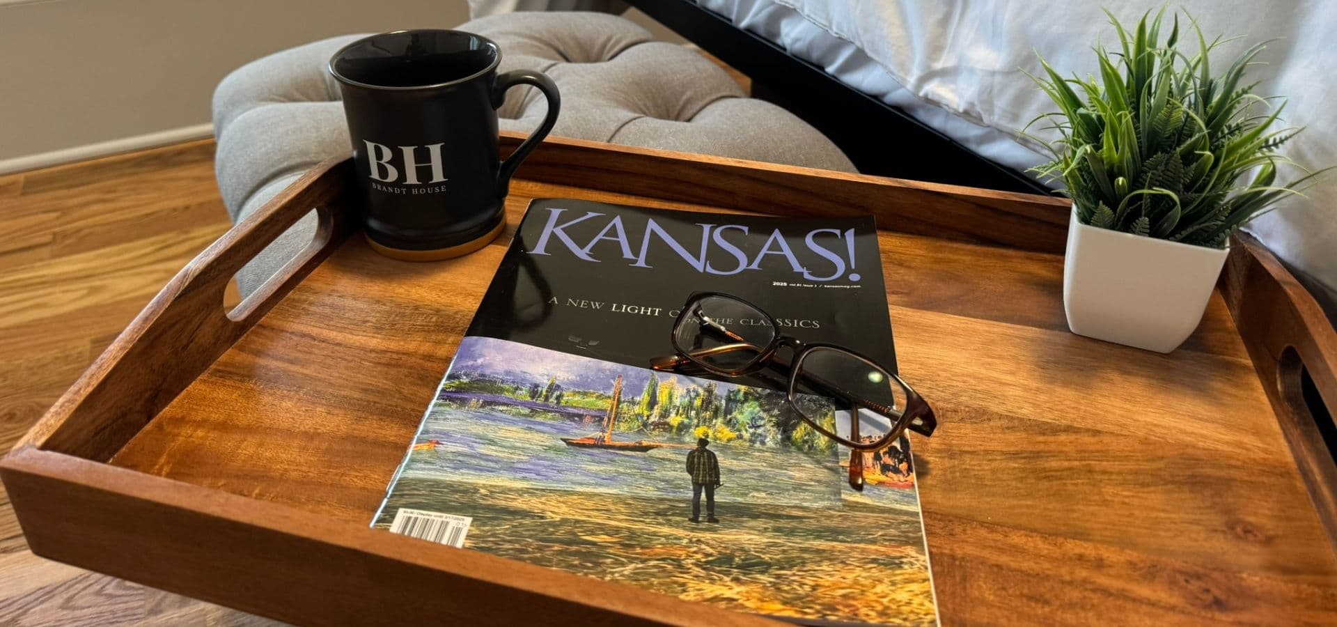 A wooden tray holds a black mug, a magazine titled "Kansas!", reading glasses, and a small potted plant.