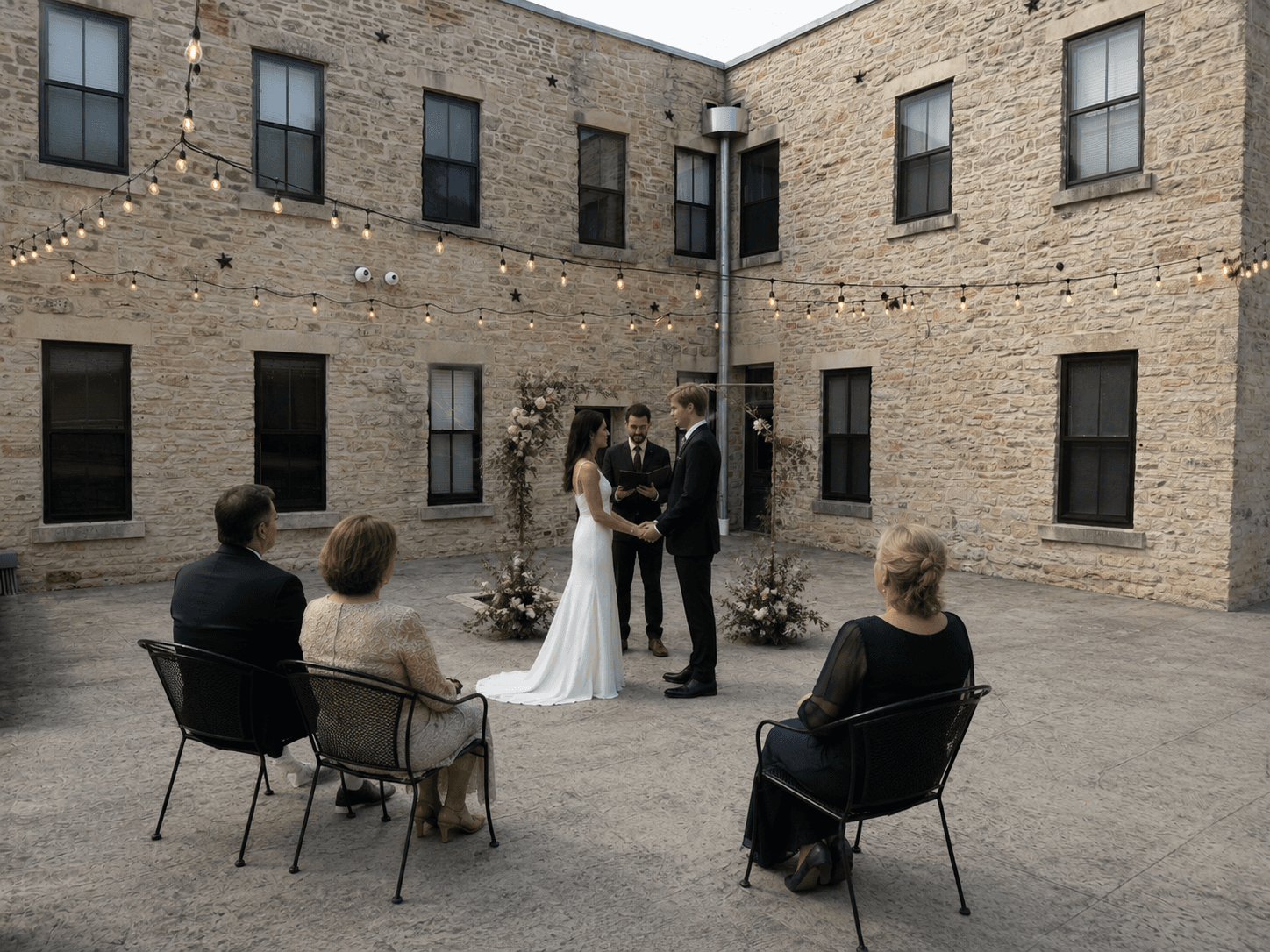 A couple exchanges vows during a wedding ceremony in a courtyard surrounded by seated guests and string lights.