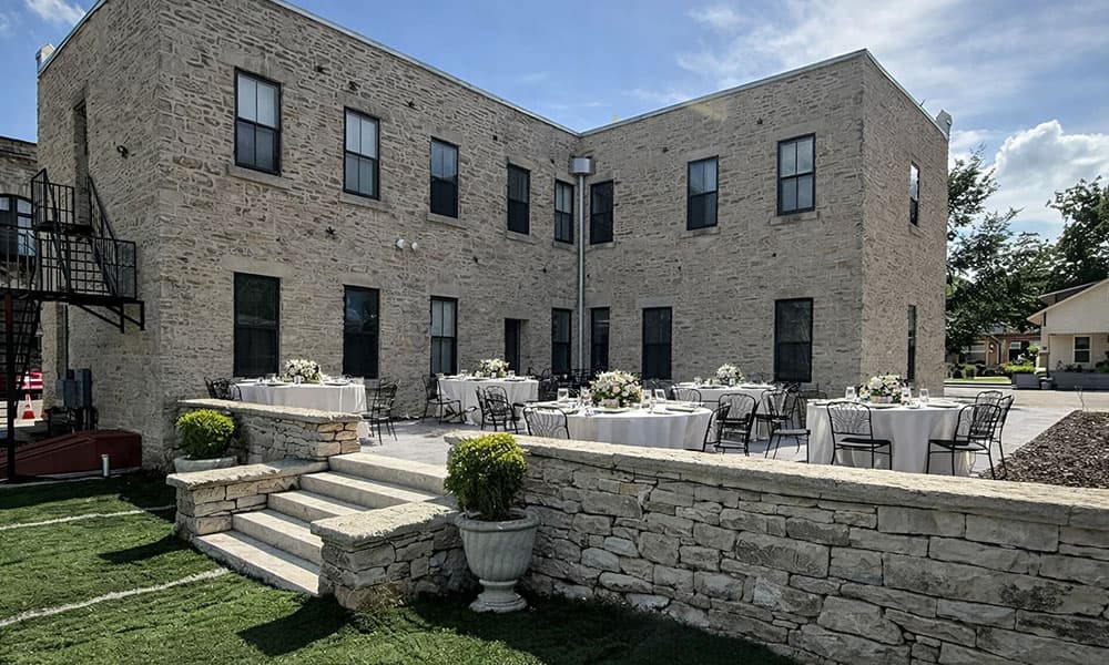 A stone building with several windows overlooks an outdoor dining area set with tables and chairs.