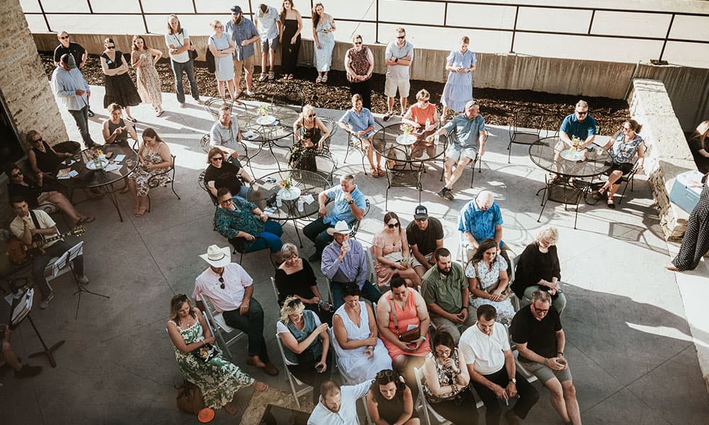 A diverse crowd of people gathers at outdoor tables, some seated and others standing, enjoying a sunny event.