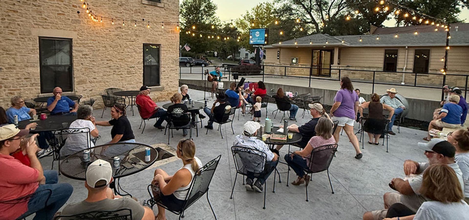 A lively gathering of people seated outdoors at tables, enjoying an evening atmosphere with string lights.