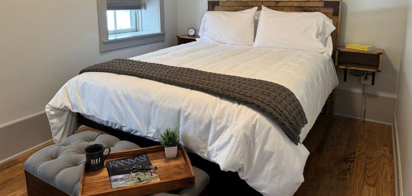 A cozy bedroom featuring a neatly made bed, a small bedside table, and a decorative stool with a coffee cup and magazine.