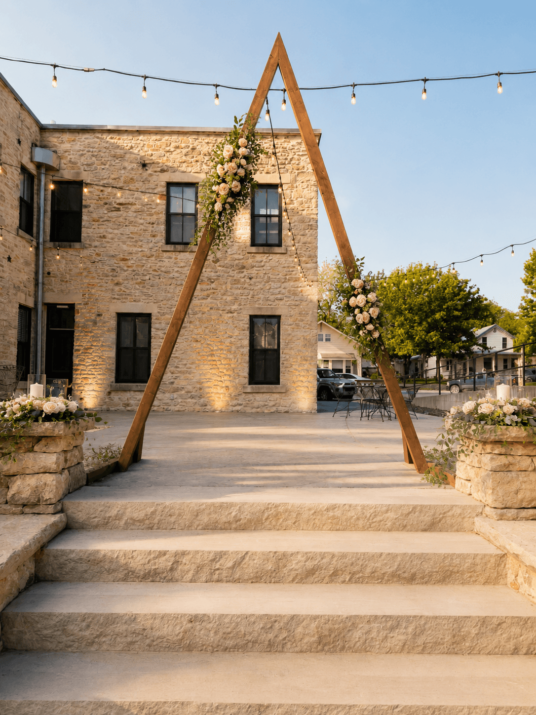 A wooden triangular arch adorned with flowers stands at the entrance of a stone building, illuminated by string lights overhead.