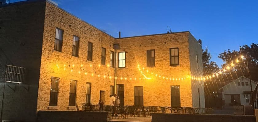 A warm-lit brick building with string lights at dusk, featuring outdoor seating.