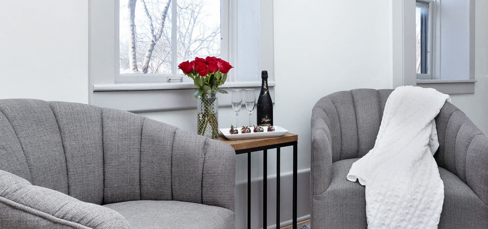 A cozy seating area featuring two gray armchairs, a small table with a bottle of champagne, glasses, chocolates, and a vase of red roses.