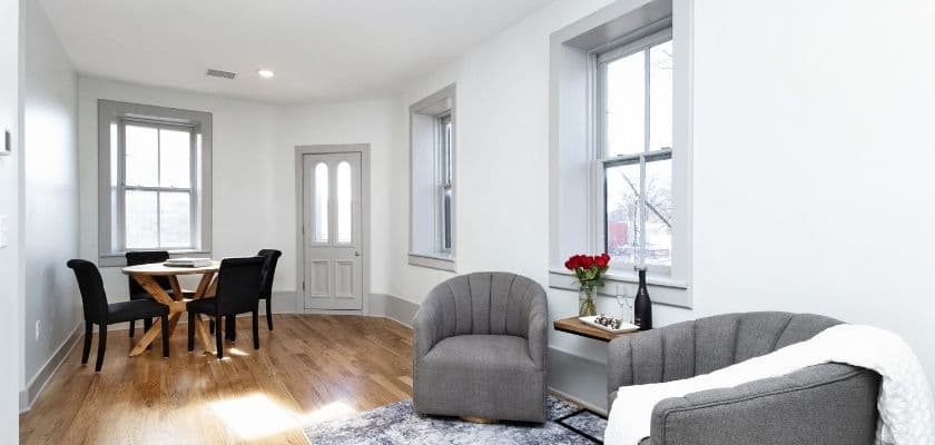 A bright guest lounge with gray chairs, a dining table, wood floors, tall windows, and simple modern decor.
