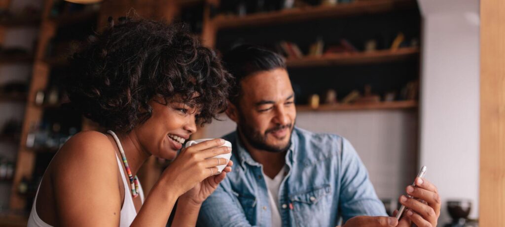 Smiling couple sitting in a cozy café, one holding a cup of coffee while they look at a smartphone together.