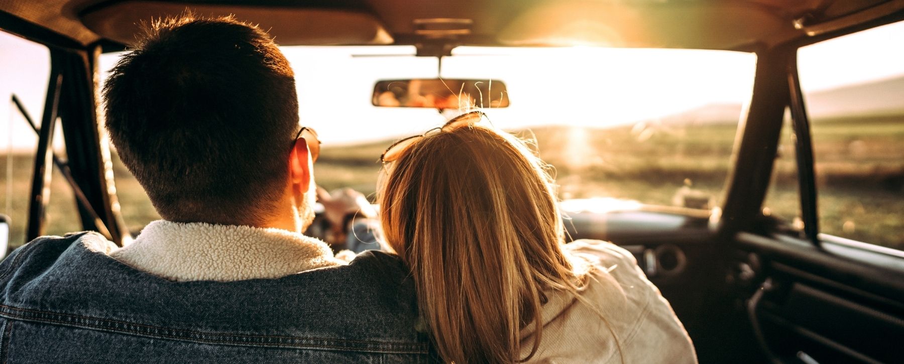 A couple sitting close together in the front seat of a car, watching the sunset through the windshield during a scenic drive.